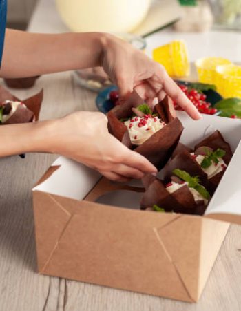 Closeup view of hands of female confectioner or baker in blue apron packing fresh decorated muffins with cream top to delivery craft box. Ready cupcakes with berries and mint in package.